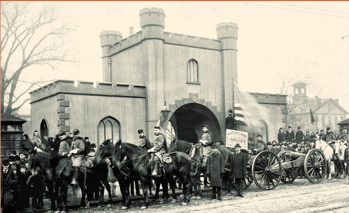 Historic photo of a parade of men and horse-mounted veterans, and at least 2 cannons on cannon carriages. An American flag waves in the parade.